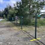 Dark green temporary mesh fencing with yellow feet is installed along a gravel path, bordering an area with tall pine trees and overgrown vegetation under a cloudy sky.