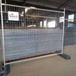 A single galvanized temporary fencing panel with welded wire mesh and dark grey plastic feet, standing in a factory setting with a stack of similar panels in the background. A white sign is attached to the top bar, representing reliable Chinese temporary fencing solutions.