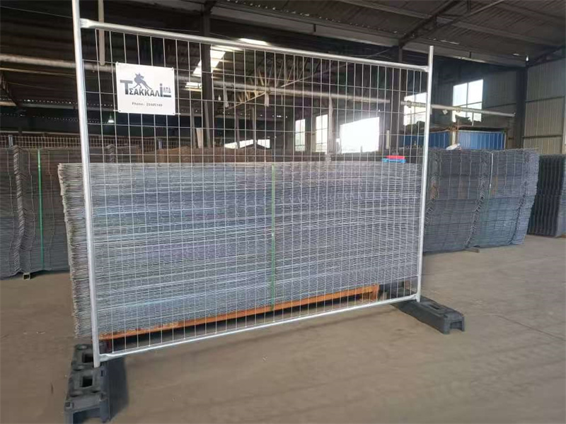 A single galvanized temporary fencing panel with welded wire mesh and dark grey plastic feet, standing in a factory setting with a stack of similar panels in the background. A white sign is attached to the top bar, representing reliable Chinese temporary fencing solutions.