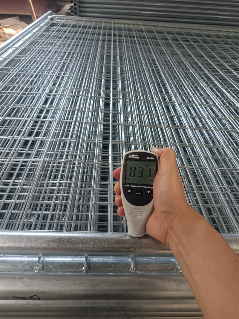 A person's hand holds a digital coating thickness gauge displaying "112" against the frame of a stacked hot-dip galvanized temporary fence panel, indicating a quality control check.