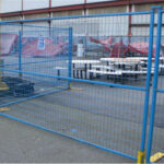 Blue temporary mesh fence panels with yellow bases are set up in an outdoor paved area, enclosing what appears to be a space with picnic tables and a large building in the background.