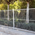 A line of galvanized temporary fencing panels with mesh infill, erected outdoors along a paved walkway with green foliage in the background, demonstrating typical event or perimeter security.