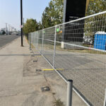 A long line of galvanized temporary mesh fencing with yellow bases extending along a sidewalk next to a road, with a portable toilet and a tall building visible in the distance under a bright sky.