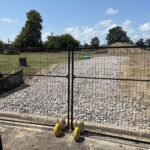 Black temporary fence double gate with integrated wheels for vehicular access, installed on a gravel path at a construction site, featuring a 'Sunbelt' sign.