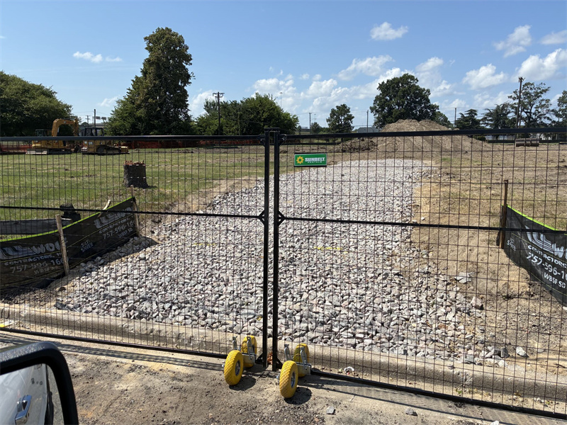 Black temporary fence double gate with integrated wheels for vehicular access, installed on a gravel path at a construction site, featuring a 'Sunbelt' sign.