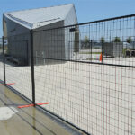 Dark grey temporary fencing with welded wire mesh panels, secured by orange plastic bases on a wet paved surface, with a modern building in the background.