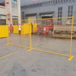 Bright yellow temporary mesh fencing with flat, sturdy bases, enclosing an area in an industrial yard with a red truck and workers visible in the background.