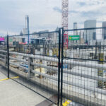 A black temporary fence with yellow bases delineates a large urban construction site, with a city skyline visible in the background.