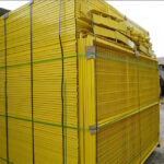 "A large stack of brightly colored yellow temporary fence panels, neatly bundled with green straps, likely prepared for shipping or storage. The panels feature a mesh design and sturdy frames, typical of construction or event barriers."