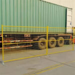 Yellow temporary fence panels with flat bases are set up on a paved surface, running in front of a large green shipping container on a truck.