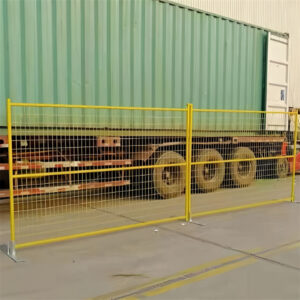 Yellow temporary fence panels with flat bases are set up on a paved surface, running in front of a large green shipping container on a truck.