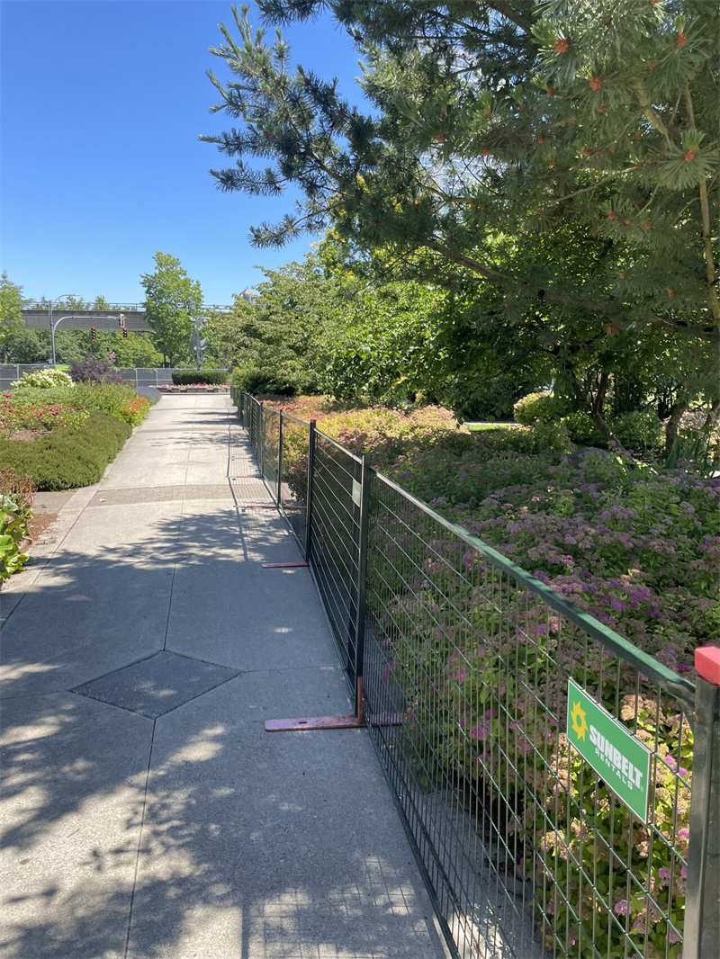 Dark green temporary fencing with red flat feet, marked with a "Sunbelt Rentals" sign, installed along a paved park pathway next to green bushes and trees under a clear blue sky.