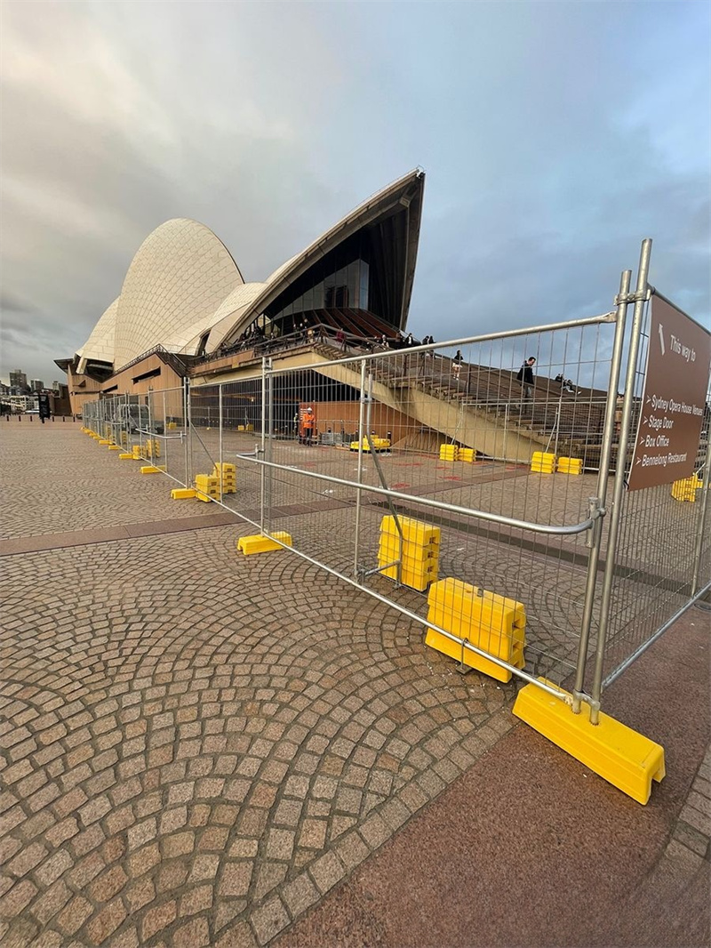 Iconic Sydney Opera House with distinctive white shell architecture surrounded by galvanized steel temporary fencing panels mounted on bright yellow plastic feet, creating restricted access area around entrance with directional signage for Stage Door, Box Office and Bennelong Restaurant visible on cobblestone forecourt under overcast sky.