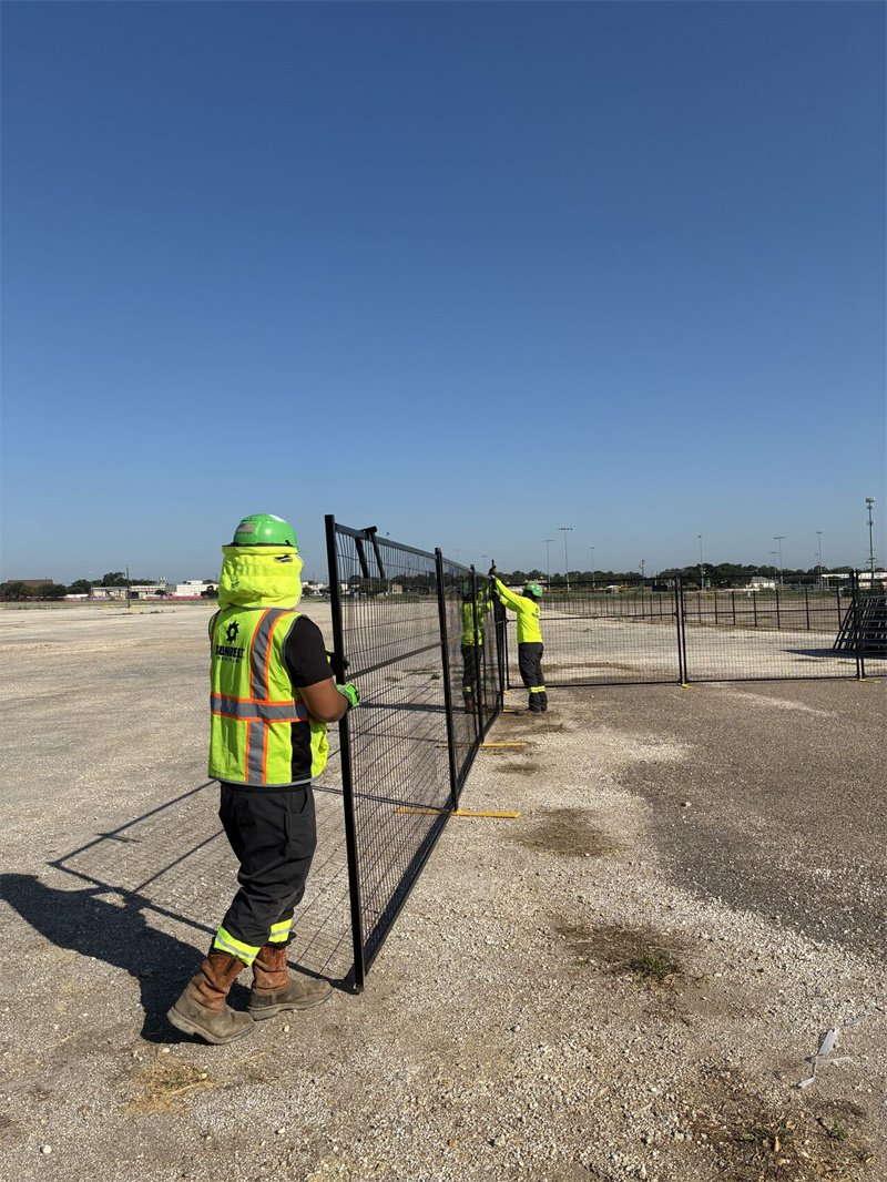 Two construction workers in high-visibility vests and hard hats are installing black temporary mesh fence panels with yellow bases on a large, open construction site under a clear blue sky.