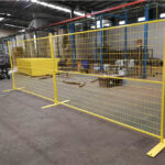 Yellow temporary fence panels with matching flat bases, arranged inside a factory or warehouse, with other yellow components and industrial equipment visible.