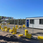 Temporary fencing with prominent yellow plastic bases enclosing an area next to a white portable building, with a billboard visible in the background under a clear sky.