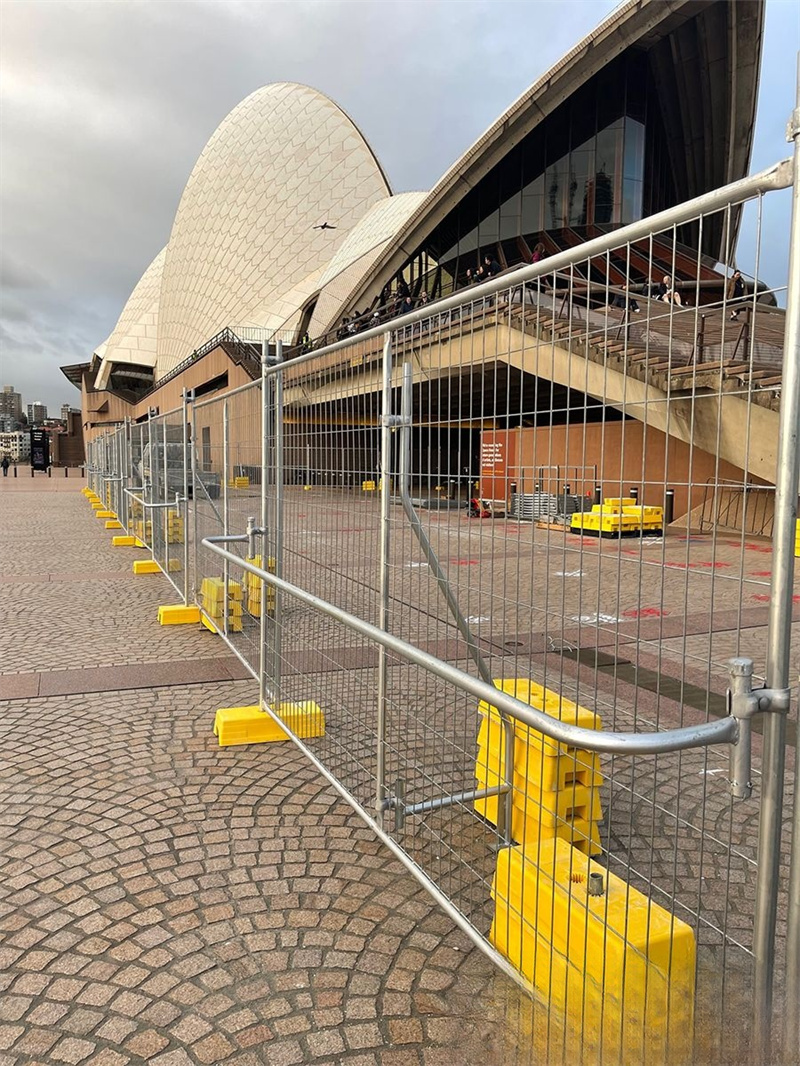 Galvanized temporary fencing with yellow plastic feet forms a barrier in the foreground, with the iconic Sydney Opera House and its distinctive white shell-like roofs visible in the background on a cloudy day.