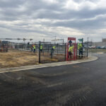 Workers in high-visibility vests are installing black temporary fencing and a red "ENTRANCE" sign around a construction site on a partially paved and dirt lot, with a street and traffic lights in the background.