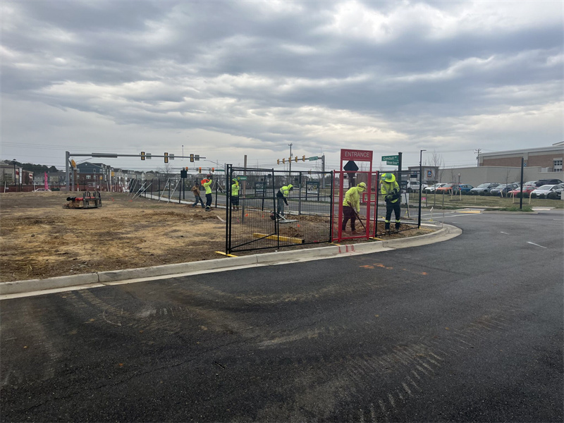 Workers in high-visibility vests are installing black temporary fencing and a red "ENTRANCE" sign around a construction site on a partially paved and dirt lot, with a street and traffic lights in the background.