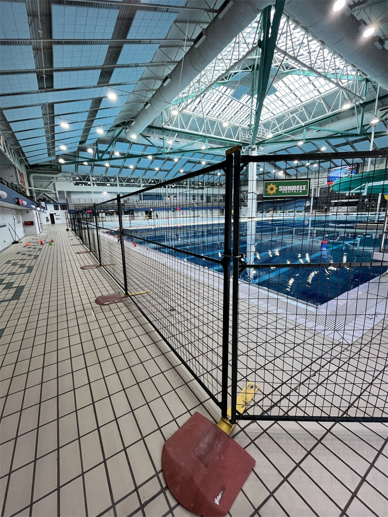 Black temporary fencing with red bases secures a section of a large indoor swimming pool facility, likely for maintenance or an event, with natural light filtering from the roof.