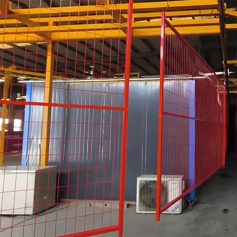 A red temporary mesh fence stands indoors in what appears to be a factory or warehouse, with industrial equipment and yellow overhead structures visible in the background, likely segmenting an area.