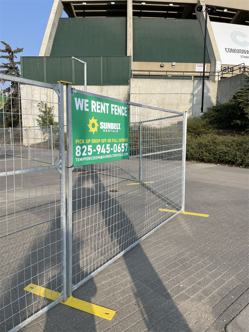 A galvanized temporary fence panel with yellow bases and a green "Sunbelt Rentals - We Rent Fence" sign, placed in front of a building with a green roof, possibly a stadium.