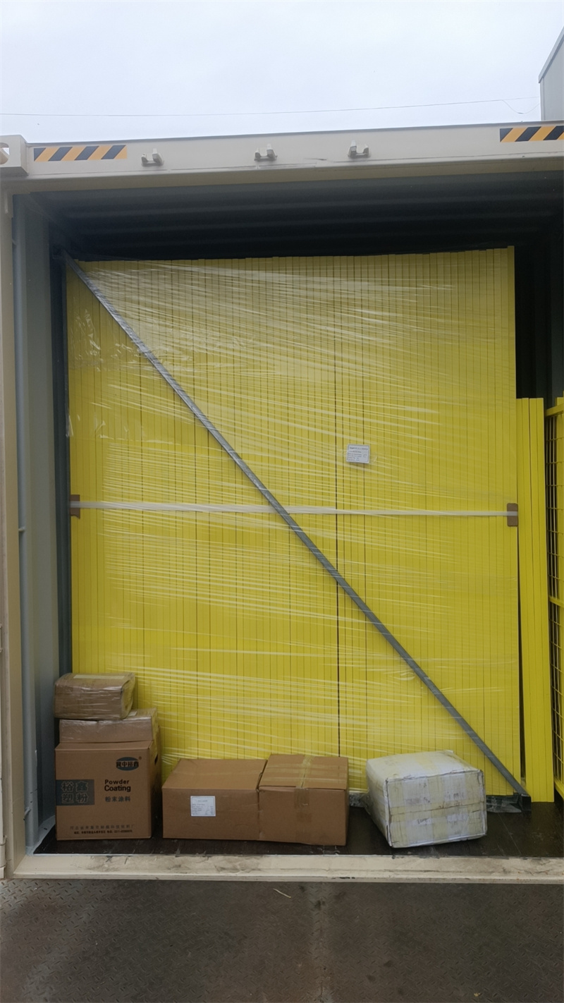 A shipping container is packed with tall stacks of yellow temporary fence panels, individually wrapped in clear plastic, alongside several cardboard boxes.