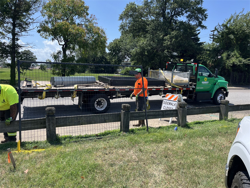 Two construction workers, wearing high-visibility vests, are handling dark temporary fence panels being transported on the back of a flatbed truck. The scene is outdoors near a paved road and a grassy area, with a "SIDEWALK CLOSED" sign visible, suggesting site preparation or removal.