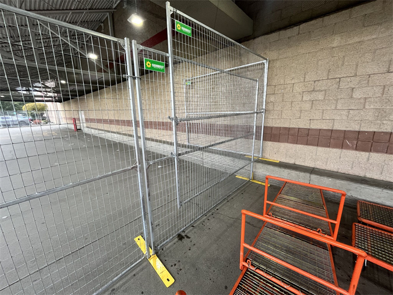 Galvanized temporary chain-link fence panels with yellow plastic bases forming a barrier in an outdoor commercial area, possibly a store entrance, with shopping carts visible.