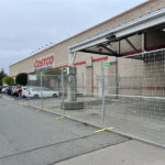 Temporary gray mesh fences with yellow bases are set up in a parking lot, blocking off an area in front of a Costco Wholesale store. Cars are parked nearby, and trees are visible in the background.