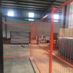 Orange temporary fence panels creating an enclosure within a warehouse, with stacked metal sheets and other materials in the background.