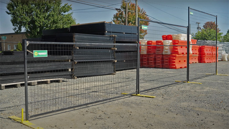 Black temporary fence panels with yellow bases, featuring a "Sunbelt Rentals" logo, separate a gravel area from stacks of construction materials and orange barriers in the background.