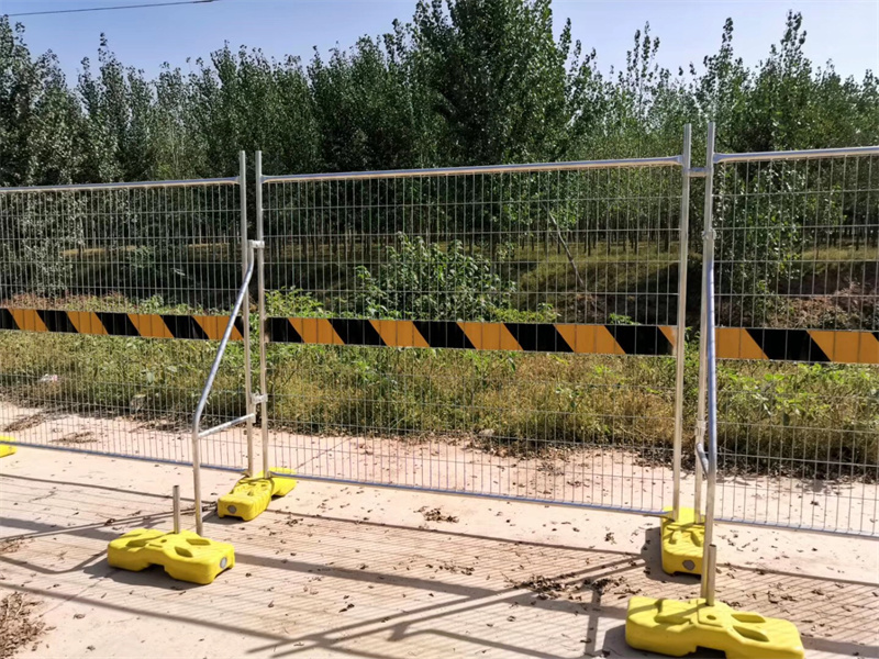 Galvanized steel mesh temporary fencing panels mounted on yellow concrete feet with black and yellow diagonal hazard warning tape, installed along paved walkway adjacent to lush green vegetation and mature trees on sunny day.