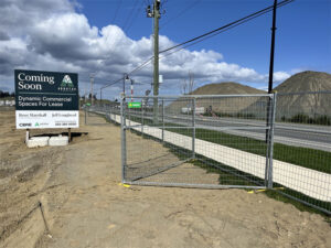 Temporary galvanized wire mesh fencing is installed along the edge of a construction or development site next to a paved road. A "Coming Soon" sign for "Dynamic Commercial Spaces For Lease" stands in the foreground, while piles of earth and gravel are visible in the background under a clear blue sky.