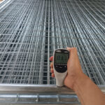 A person's hand holds a digital coating thickness gauge displaying "112" against the frame of a stacked hot-dip galvanized temporary fence panel, indicating a quality control check.