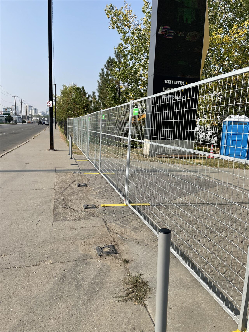 A long line of galvanized temporary fencing with yellow bases stretches along a paved sidewalk next to a road, with a blue portable toilet visible behind the fence.