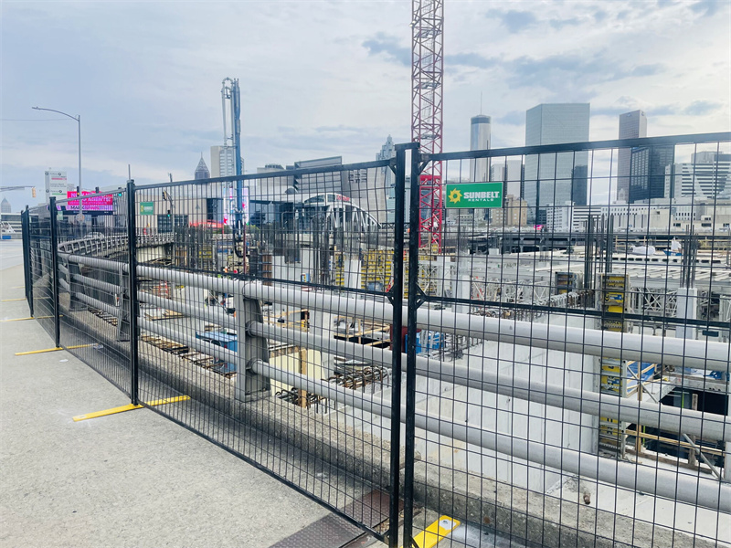 A black temporary fence with yellow bases delineates a large urban construction site, with a city skyline visible in the background.
