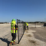 Two construction workers in high-visibility gear are setting up black temporary fence panels in a large, open dirt field under a bright blue sky.