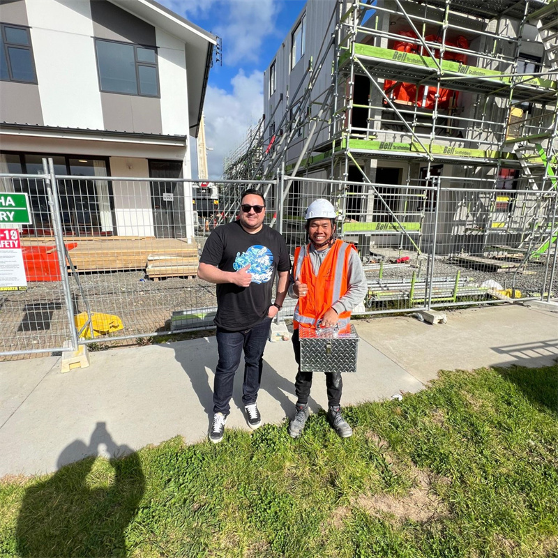 Two men, one in a safety vest and hard hat holding a toolbox, and the other in a t-shirt, stand smiling and giving thumbs-up in front of temporary fencing at a residential construction site with new buildings and scaffolding.