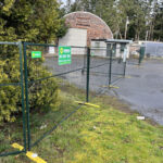 Green-framed temporary welded mesh fencing with yellow bases, including a gate section with wheels, installed on a grassy area next to an asphalt path. A "SUNBELT WE RENT FENCE" sign is attached to the gate, and a distinctive round building with a wooden shingle roof is visible in the background among trees.