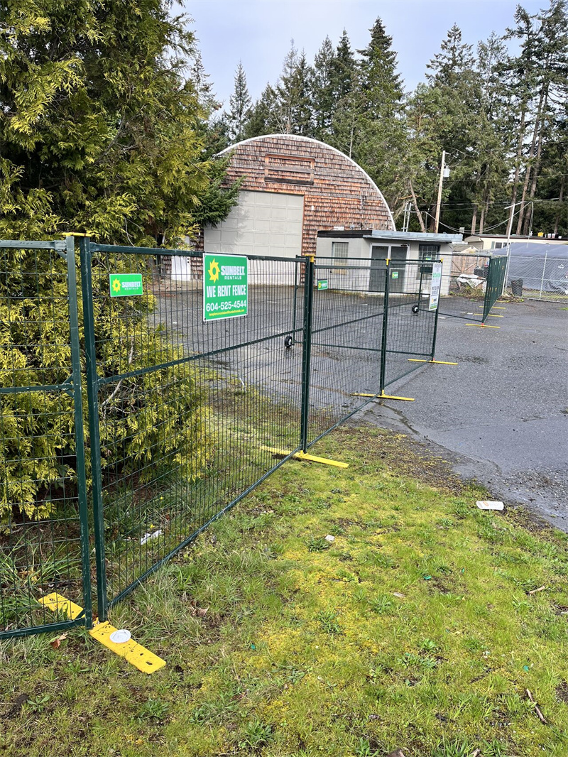 Green-framed temporary welded mesh fencing with yellow bases, including a gate section with wheels, installed on a grassy area next to an asphalt path. A "SUNBELT WE RENT FENCE" sign is attached to the gate, and a distinctive round building with a wooden shingle roof is visible in the background among trees.