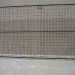 A single black temporary fence panel with orange flat bases, positioned against a plain light gray wall.