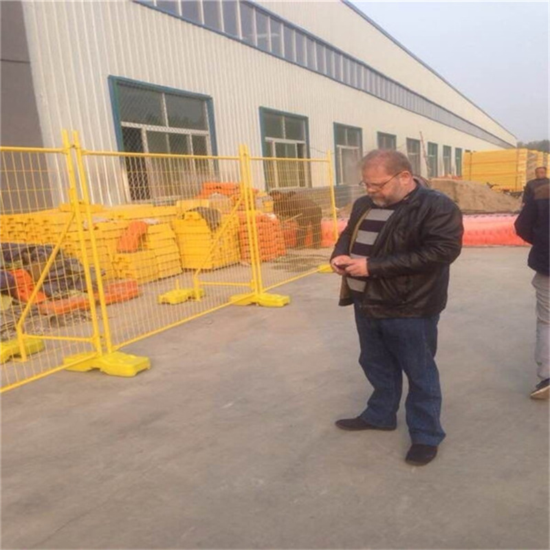 Quality control inspector in black leather jacket examining yellow powder-coated temporary fencing panels with matching plastic bases installed outside large industrial manufacturing building with multiple windows.