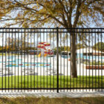 Black vertical picket fence providing a clear view into a vibrant outdoor water park with colorful slides and splash pads, illustrating commercial pool safety fencing.
