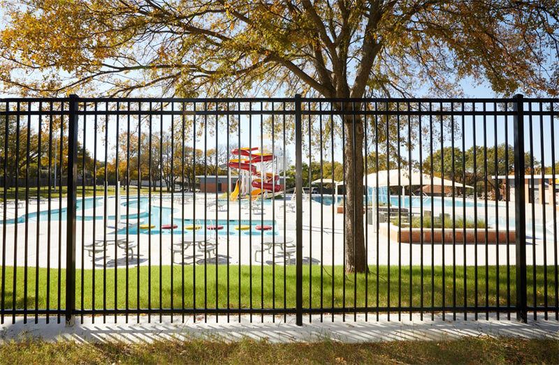 Black vertical picket fence providing a clear view into a vibrant outdoor water park with colorful slides and splash pads, illustrating commercial pool safety fencing.