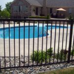 A black vertical picket fence surrounding an in-ground swimming pool in a landscaped residential backyard, with a large house visible in the background.
