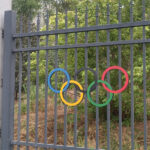 A close-up of the Olympic rings attached to a gray steel picket fence with foliage in the background.
