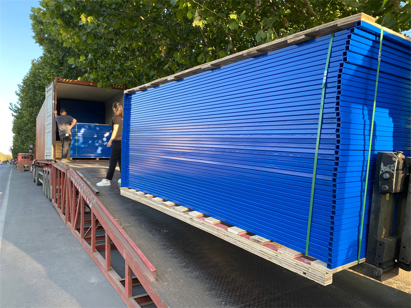 A large quantity of blue temporary fence panels, neatly stacked on wooden pallets, being loaded onto a semi-trailer truck for shipping.