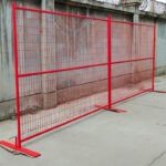 Two red temporary fence panels with welded mesh and flat steel base plates, installed outdoors against a concrete wall with barbed wire for security.