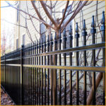 A black ornamental steel fence with decorative finials installed in front of a house with beige siding and a bare tree.